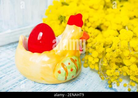 Osterkomposition. Ein rot bemaltes Ei in einem Keramik-Huhn mit einem Bouquet von frühlingsgelben Mimosen auf dem Ostertisch. Stockfoto