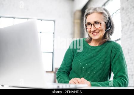 Fröhliche alte Frau mit Headset, Falten im Gesicht, lächeln, hören am Computer, Beratung des Kunden online, gibt den Rat, Heimarbeit, positive Person, nimmt jede Chance zu arbeiten Stockfoto