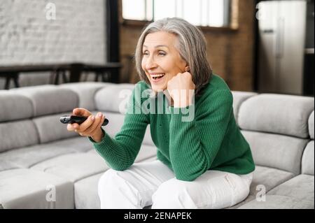 Bild von emotionalem Lächeln optimistische gealterte Frau grauhaarig, gemütlich gekleidet, sitzen auf einem bequemen Sofa mit einer Fernbedienung in der Hand, sehen lustige Programm im Fernsehen oder aufgeregt Film, Zeit zu Hause verbringen Stockfoto