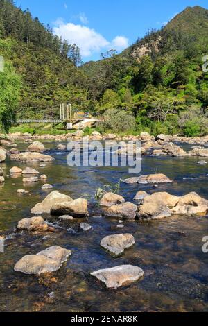 Der felsige Ohinemuri River fließt durch die Karangahake Gorge, Neuseeland. Im Hintergrund überquert eine Hängebrücke den Fluss Stockfoto