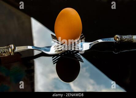 Frisches Hühnerei (Hühnerei) auf einer Zusammensetzung aus zwei miteinander verflochtenen Gabeln über schwarzem Boden ausgeglichen. Selektiver Fokus. Stockfoto