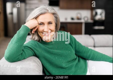 Lächelnde ältere Frau mit grauem, gepflegtem Haar, blauen Augen und schönen Falten, die zu Hause auf dem Sofa sitzt, den Kopf auf einer Hand ruht und die Kamera anschaut, fühlt sich ruhig und ausgeglichen, freundlich an Stockfoto
