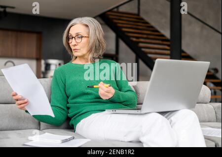 Serious ältere Geschäftsfrau konzentriert während der Prüfung von Dokumenten, sitzen auf dem Wohnzimmer, Laptop auf dem Schoß, freiberuflicher Anwalt, Vorbereitung einer Rede, Home Office, Beratung Client online Stockfoto