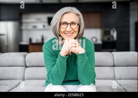Gesunde ältere Frau mit grauen Haaren mit leuchtend blauen Augen, elegante Brille, sitzt auf dem reinen bequemen Sofa in der modernen Wohnung, in grünen Pullover und weißen Hosen gekleidet, legte Kinn auf die Hände Stockfoto