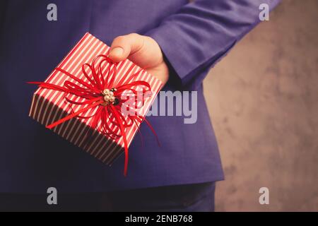 Geschäftsmann Mann in Anzug hält Geschenk hinter seinem Rücken. Rote Box mit Bändern in der Hand verziert. Mockup, Platz zum Schreiben auf grauem Hintergrund. Stockfoto