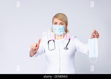 Ein Arzt in einem medizinischen Kleid, eine Frau mit schützender medizinischer Maske auf ihrem Gesicht zeigt Zeigefinger nach oben für Aufmerksamkeit Zeichen, hält einen medizinischen Einweg Stockfoto