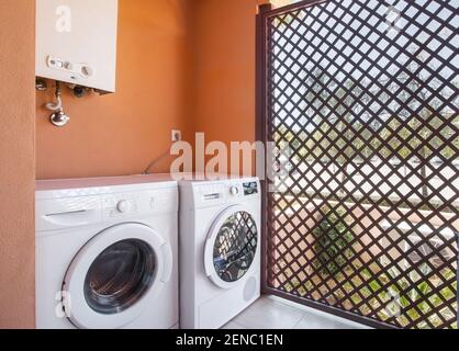 Einfache Waschküche mit einer Waschmaschine zum Waschen und Trocknen, einem Kessel auf der Oberseite und einer eingezäunten Teilung, die den Garten im Freien zeigt. Stockfoto