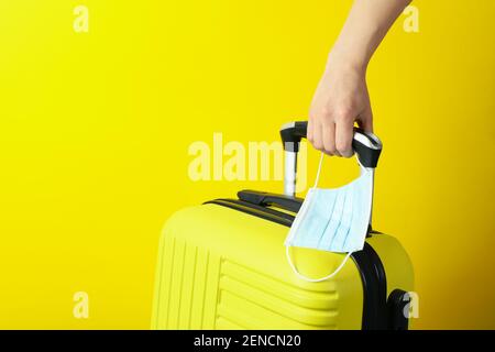 Weibliche Hand halten Reisetasche und medizinische Maske auf gelb Hintergrund Stockfoto