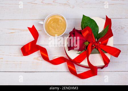 Tasse Kaffee und herzförmige Geschenkbox mit rotem Band und roter Rose auf weißem Holzhintergrund. Valentinstag romantische Wohnung Lay Top View Frühstück Stockfoto