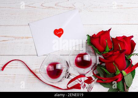 Weingläser mit Rotwein, Umschlag und roten Rosen mit rotem Band auf weißem Holzhintergrund. Valentinstag romantisches Abendessen flat Lay Top View Konzept Stockfoto