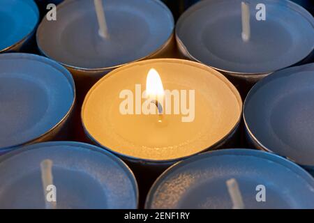 Nahaufnahme von Teelampen mit einer beleuchtet. Eine einzelne warme orange Flamme zwischen Kerzen im blauen Schatten. Weihnachten, Beileid oder Begräbnishintergrund. Stockfoto