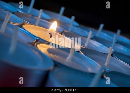 Nahaufnahme von Teelampen mit einer beleuchtet. Eine einzelne warme orange Flamme zwischen Kerzen im blauen Schatten. Weihnachten, Beileid oder Begräbnishintergrund. Stockfoto