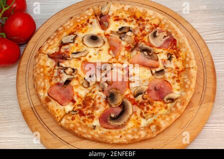 Pizza mit Schinken, Pilzen und Käse auf einem Brett mit Tomaten Stockfoto