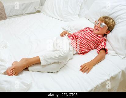 Porträt eines lächelnden Babys im karierten Hemd und mit Sonnenbrille auf einem weißen Bett. Im Urlaub an einem Sommertag. Stockfoto