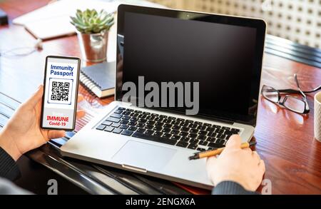 Immune Coronavirus digitalen Gesundheits-Pass auf Handy, Mann mit einem leeren Bildschirm Laptop, Büro Hintergrund, Covid19 Impfung und Flug Stockfoto