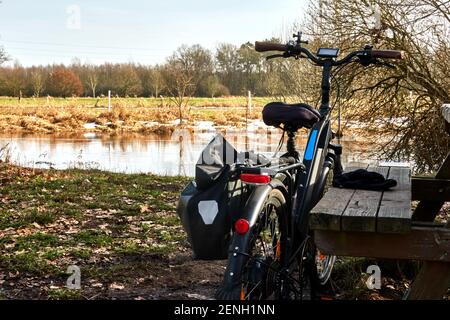 Schwarzes Fahrrad mit Gepäcktasche an einer rustikalen Ruhe gelehnt Bank vor einem kleinen Fluss in der Heide Querformat Stockfoto
