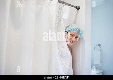 Eine Frau lächelt, als sie mit einer Duschhaube aus einer Duschkabine gucken kann. Stockfoto