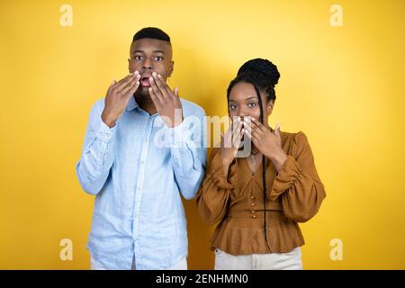 Junges afroamerikanisches Paar, das über gelbem Hintergrund steht, schockiert, den Mund mit Händen für Fehler zu bedecken. Geheimes Konzept. Stockfoto