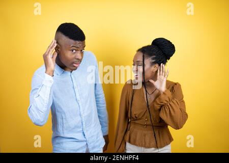 Junges afroamerikanisches Paar, das über gelbem Hintergrund steht, überrascht mit Überreichen Sie das Ohr und hören Sie Gerüchten oder Klatsch zu Stockfoto
