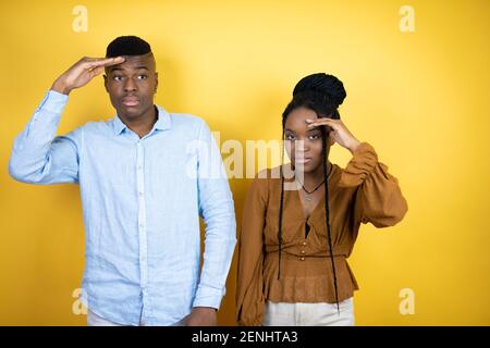 Junges afroamerikanisches Paar, das über gelbem Hintergrund sehr glücklich und lächelnd steht und weit weg mit Hand über Kopf schaut. Suchkonzept. Stockfoto