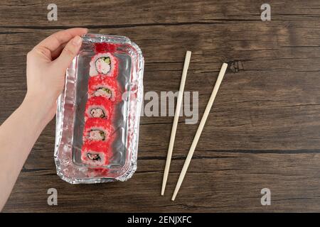 Weibliche Hände halten Aluminiumfolie Platte von Sushi Rollen auf Holzoberfläche Stockfoto