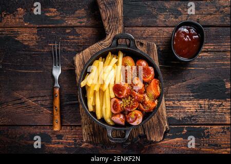 Currywurst Street Food Mahlzeit, Curry Gewürz auf Würsten serviert Pommes in einer Pfanne. Dunkler Holzhintergrund. Draufsicht Stockfoto