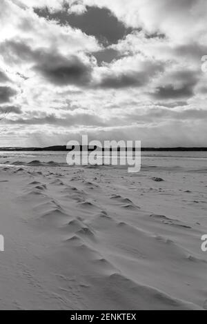 Schwarz-Weiß-Foto von Mustern im Schnee am Ufer der Grand Traverse Bay mit Power Island in der Ferne unter einem bewölkten Himmel. Stockfoto