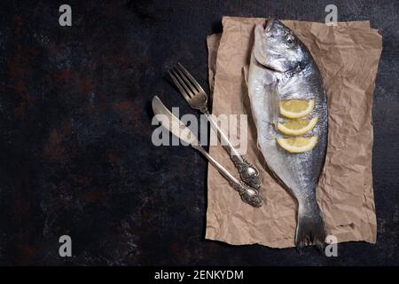 Frischer dorado Fisch auf Papier mit Besteck. Auf dunklem Grund. Kochen mit Olivenöl, Zitrone, Chili, Knoblauch, Salz, Pfeffer. Gericht des Mittelmeers d Stockfoto