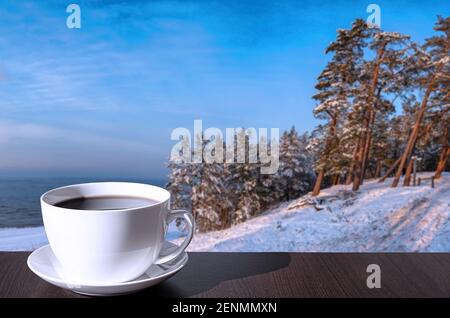 Tasse Kaffee auf dem Tisch mit Blick auf schneebedeckten Strand, Ostsee und bedeckt mit Schnee Tannen und Pinien auf dem Hügel. Stockfoto