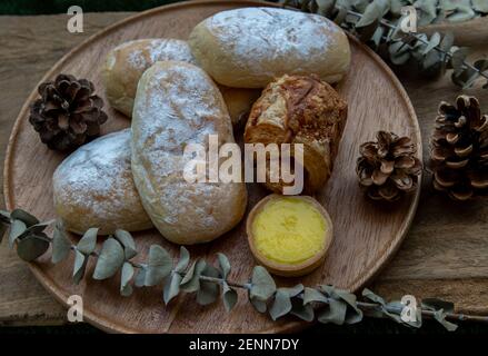 Hausgemachte süße Sahne-Softbrötchen, gebackene Croissant-Wurst und Zitronentarte mit getrockneten Blumen auf runden Holzteller auf rustikalem Holzboden. Bäckerei und d Stockfoto