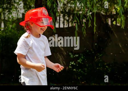 Ein kleines Mädchen im jungen Schulalter, das einen roten Feuerwehrmann-Spielzeughelm trägt, der mit Wasser besprüht wird, im Freien, Kind, das draußen spielt, Portrait zukünftiger Besetzer Stockfoto