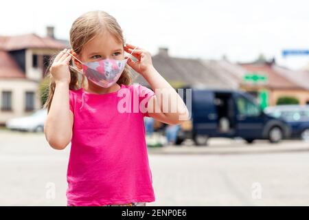 Kleines Mädchen, ein Kind im Schulalter, das eine farbenfrohe moderne Schutzmaske an- oder auszieht, Outdoor Lifestyle potrait, Stadtgebiet, Kopierraum. Stockfoto