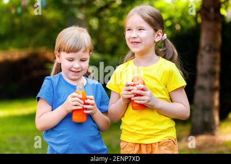 Zwei glückliche junge fröhliche kleine Mädchen im Schulalter halten geöffnete Glasflaschen voll von gesunden Orangensaft Obst trinken lächelnd, im Freien portra Stockfoto