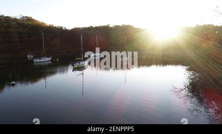 Die Sonne platzt durch die Bäume über einem ruhigen See Und eine Auswahl an Segelbooten Stockfoto