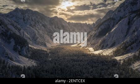 Luftflug über das Bergtal im Winter mit Schnee bedeckt. Filmische Drohnenansicht mit Sonnenlicht, das durch Wolken bricht. Stockfoto