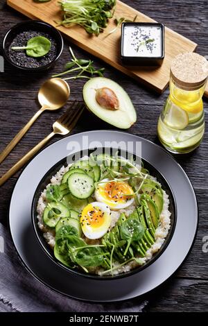 Quinoa Schüssel mit Ei gekrönt mit Sonnenblumen-Microgreens, Gurke, Avocado, Baby Spinat in einer schwarzen Schüssel auf einem dunklen Holztisch, türkische Küche, vertikale V Stockfoto