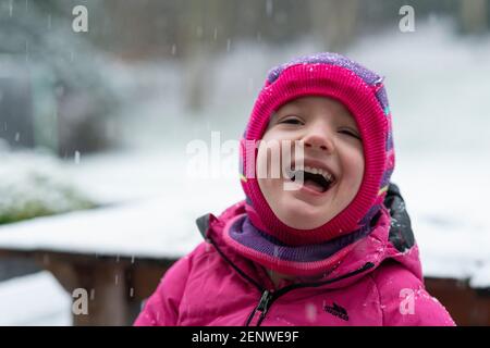 Kleines Mädchen lachen und genießen Sie den Schnee im Garten in Großbritannien, rosa Hut und rosa Mantel, Winterspaß im Garten, Schnee Spaß, Stockfoto