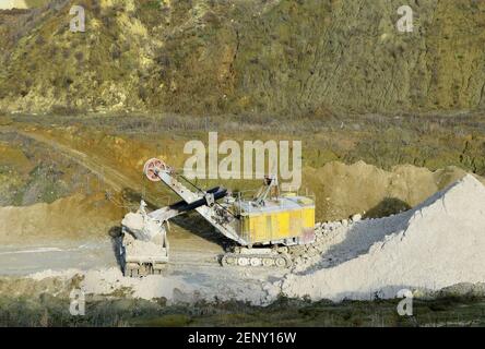 Bagger im Steinbruch lädt Gestein in einen Bergbauwagen. Bagger und Lastwagen arbeiten in einer Kreidegrube. Luftaufnahme der Bergschlucht mit Bergen und Stockfoto