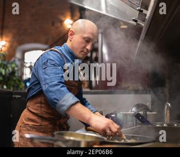 Glatze Mann in Schürze braten leckeres Essen auf Pfanne, während Arbeiten in der Küche des Restaurants Stockfoto