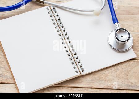 Leeres Notizbuch aus Papier und blaues Stethoskop auf braunem Holztischhintergrund. Draufsicht mit Kopierbereich (selektiver Fokus). Büroschreibtisch Konzept mit Stockfoto