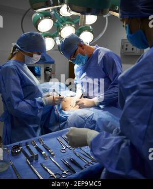 Operationsteam in sterilen Handschuhen und Schutzmasken tun ästhetische Chirurgie im Operationssaal. Assistent steht neben OP-Tisch mit medizinischen Werkzeugen. Konzept der Medizin und plastische Chirurgie. Stockfoto