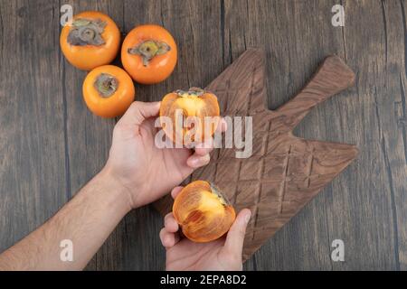 Männliche Hände halten halbierte Kaki Früchte auf Holzoberfläche Stockfoto