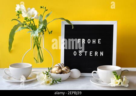 Text Frohe Ostern bedeutet Frohe Ostern. Briefbrett mit Frühling Freesia Blumen und Tee-Tasse auf dem Tisch. Schwarzer Briefbogen mit Text. Natürliche Ostereier, Stockfoto