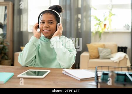Nette Schülerin der afrikanischen Ethnizität in Kopfhörer Blick auf Sie Beim Sitzen am Tisch im Wohnzimmer gegen großes Fenster und Musik hören Stockfoto