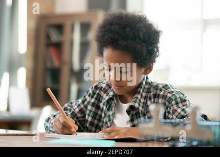 Nette ernsthafte Schuljunge der afrikanischen Ethnie macht Notizen im Copybook Während Hausaufgaben am Tisch im Wohnzimmer gegen Computer Stockfoto