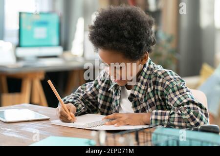 Elementare Schuljunge der afrikanischen Ethnizität macht Notizen in Copybook, während Hausaufgaben am Tisch im Wohnzimmer gegen Computer Stockfoto