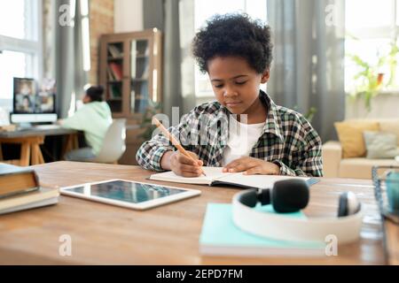 Nette elementare afrikanische Schuljunge mit Bleistift macht Notizen im Copybook Während Hausaufgaben am Tisch im Wohnzimmer gegen seine Schwester Stockfoto