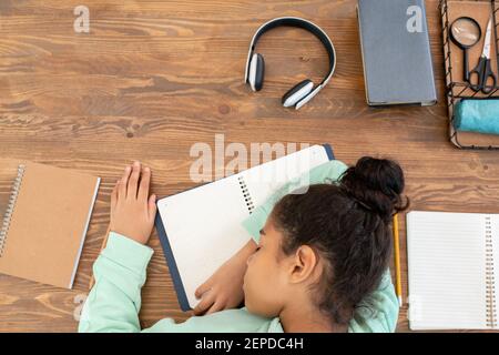 Überblick über müde gemischt Rennen Schülerin halten ihren Kopf auf offen Das Copybook auf dem hölzernen Tisch während der Vorbereitung der Hausaufgaben Stockfoto