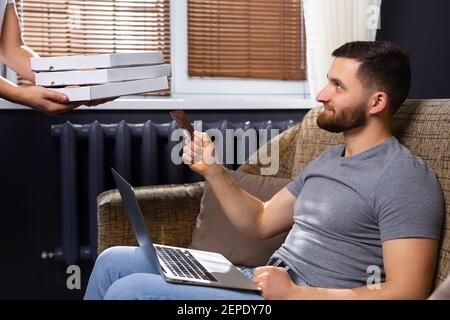 Kurier Lieferung von Pizza für einen jungen Mann, der von zu Hause an einem Remote-Job arbeitet. Zahlung per Kreditkarte. Notizbuch. Online arbeiten. Stockfoto