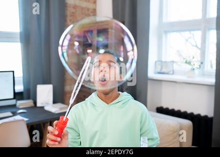 Nettes Mädchen im elementaren Alter in Casualwear weht riesige Seife Bubble, während Sie vor der Kamera stehen und Spaß haben In der häuslichen Umgebung Stockfoto